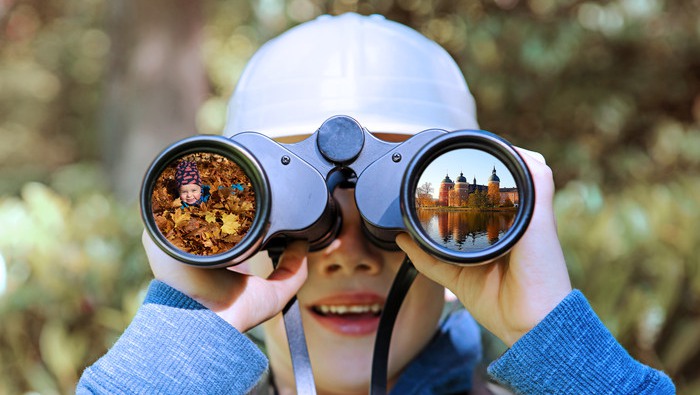 This is a close-up portrait image of a little child who is looking through big binoculars towards the camera. The boy is here seen outdoors in summer and he is wearing a pith helmet and holding binoculars straight towards the camera in such a way that his face is almsot completely covered.