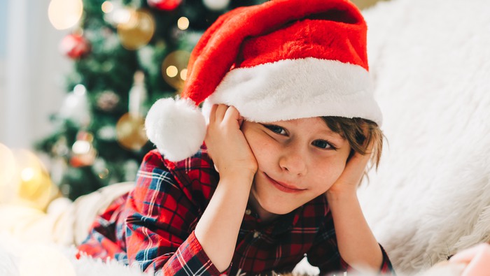 Merry Christmas. Funny Thoughtful little child lying on couch with blanket at winter holidays. Kid boy in Santa hat. Christmas tree with light bokeh. Happy new year.